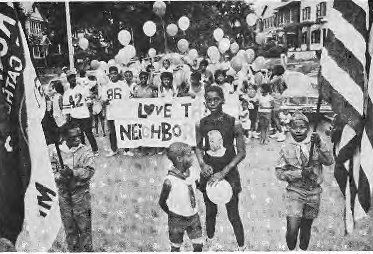 Children and neighbors carrying flags, balloons and Love Thy Neighborhood sign.